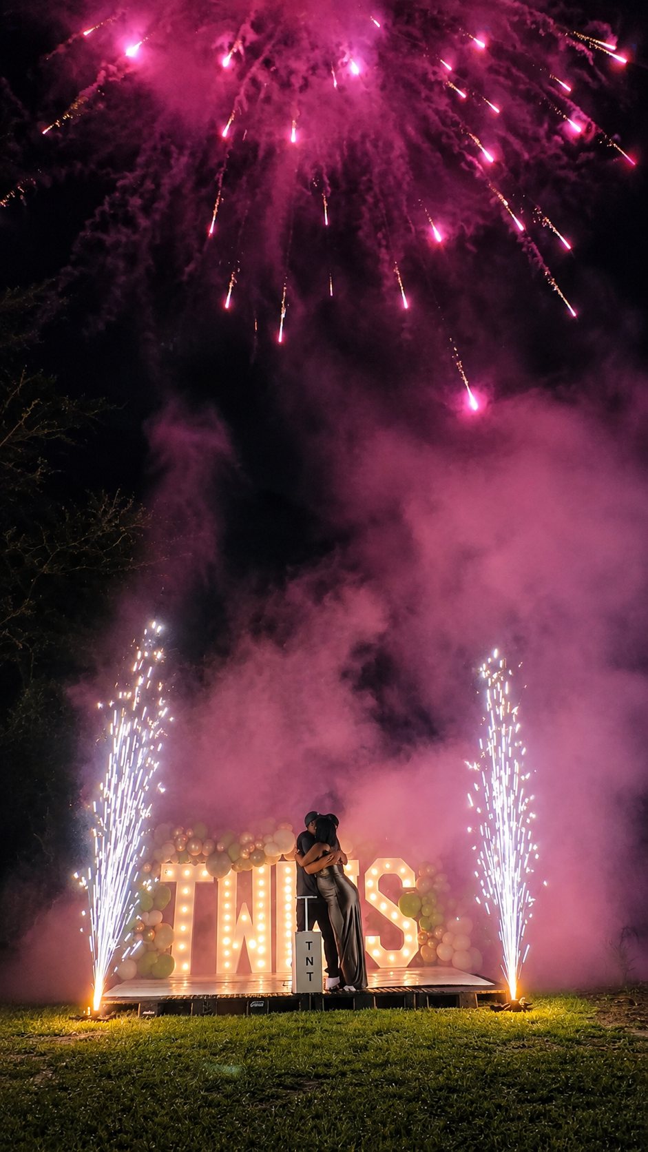 A couple embracing under pink fireworks beside glowing TWINS marquee letters at their twins gender reveal